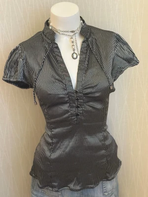 Randig svartvit blus med puffärm - Snygg svartvit blus med smala vertikala ränder och puffiga korta ärmar. Blusen har en liten ståkrage, djup v-ringning med dekorativa knappar och figurnära passform. Materialet är glansigt och ger en elegant look. Perfekt att styla med jeans eller kjol.