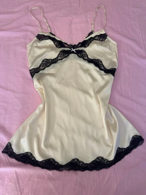 Krämfärgat nattlinne med spetsdetaljer - Sött nattlinne i krämfärgad satin med tunna axelband och svarta spetsdetaljer längs byst, midja och nederkant. Liten rosett framtill för extra detalj. Materialet känns mjukt och glansigt, perfekt för en lyxig känsla på kvällen.