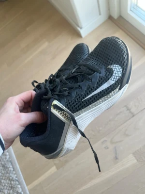 Svarta träningsskor från Nike  - Snygga svarta Metcon 6 från Nike med vit sula och mesh-överdel för bra ventilation. Skorna har ett coolt rutigt mönster på swoosh-loggan och detaljer i vitt och grått. Klassisk snörning och låg profil gör dem perfekta för en sportig look.