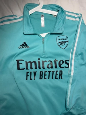 Arsenal FC x Adidas Training Top - Storlek XL (Passar även L/M) - Snygg Arsenal-träningsjacka i bra skick!! Säljer en officiell 1/4 zip träningsjacka från Arsenal FC i en fräsch turkos/mintgrön färg. Jackan har Adidas ikoniska ränder och broderade loggor. • Märke: Adidas (Original) • Storlek: Märkt XL, men då det är en slim-modell passar den utmärkt även dig som bär Large, eller som en snygg "oversized" passform för dig med Medium. • Egenskaper: AEROREADY-material som andas, tumhål i ärmarna för extra komfort. • Skick: Toppskick! Inga fläckar, hål eller slitag