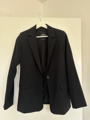 Svart kavaj från Pull&Bear - Stilren svart kavaj från Pull&Bear med klassisk passform och en knapp framtill. Kavajen har två fickor. Kavajen har en clean och modern look. Materialet är slätt och fodrat för extra komfort - perfekt till varmare väder! ☀️