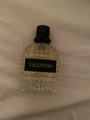 Valentino parfym med nitar - Säljer en lyxig parfym från Valentino i en transparent glasflaska med nitar och svart etikett med guldfärgad text. Flaskan har en unik, mönstrad design och svart kork som ger en exklusiv känsla.