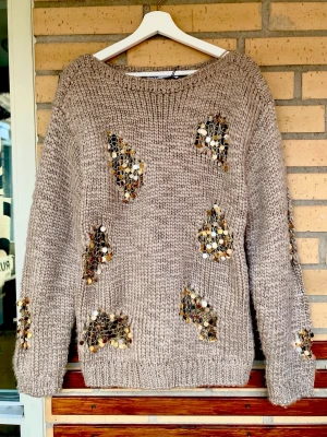 Beige stickad tröja med paljetter - Säljer en chunky beige stickad tröja med stora paljettdekorationer i guld och silver på fram- och ärmar. Tröjan har rund halsringning, lång ärm och ribbade muddar. Perfekt för dig som vill sticka ut lite extra med glittriga detaljer.