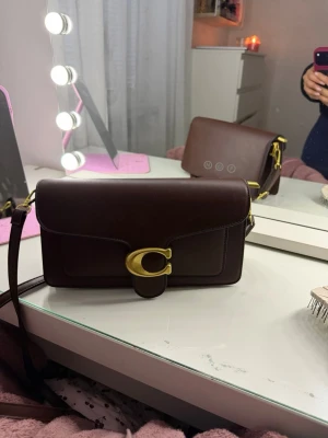 Mörkbrun handväska i läder - Snygg mörkbrun handväska  i slätt läder med axelrem. Väskan har ett stort guldfärgat C-spänne framtill och flera fack inuti, inklusive ett med dragkedja. Perfekt för dig som gillar klassisk och stilren design.