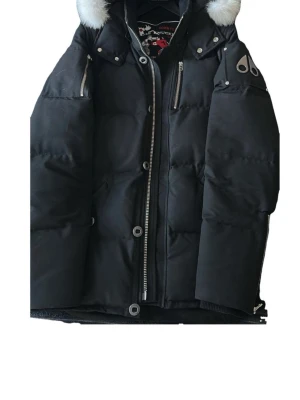 Svart dunjacka Moose Knuckles - Snygg svart dunjacka från Moose Knuckles med avtagbar huva och vit fuskpäls. Jackan har flera dragkedjeförsedda fickor, logotyp på ärmen och broderad hockeypatch på insidan. Perfekt för kalla vinterdagar och riktigt varm.