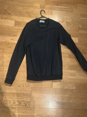 Svart sweatshirt från Stone Island - Säljer en svart sweatshirt från Stone Island med klassisk rund hals och ribbade muddar. Tröjan har den ikoniska Stone Island-loggan på vänster ärm. Perfekt för dig som gillar streetwear och vill ha en stilren look. Har hål i änden av vänstra ärmen, men inget svårt att fixa