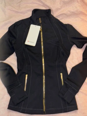 Svart jacka från Lululemon med guldzip - Snygg svart jacka från Lululemon med guldfärgade dragkedjor framtill och på fickorna. Figurnära passform och hög krage ger en sportig look. Tillverkad i stretchigt syntetmaterial som passar perfekt till vardags eller träning.