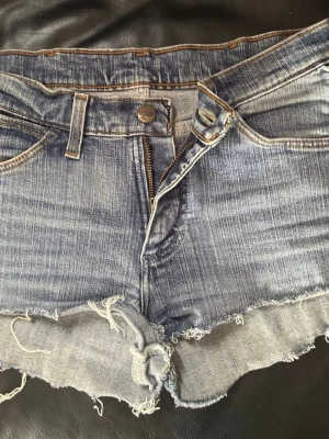 Blå jeansshorts från Wrangler - Säljer ett par klassiska blå jeansshorts från Wrangler med råa, fransiga kanter och femficksmodell. Shortsen har normal passform, bälteshällor och dragkedja framtill. Wrangler-logga på bakfickan. Perfekta för sommaren!