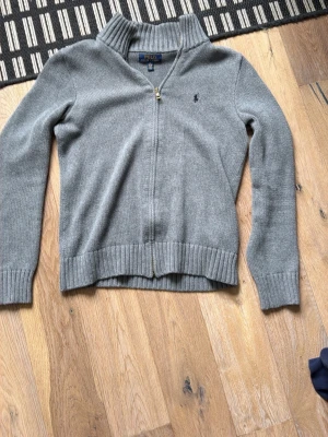 Grå ziptröja från Polo Ralph Lauren - Snygg grå ziptröja från Polo Ralph Lauren med ribbade muddar och hög krage. Tröjan har dragkedja framtill och den klassiska lilla loggan broderad på bröstet. Perfekt för lager på lager och enkel att matcha med det mesta.