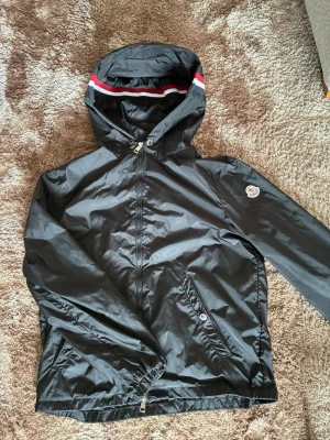 Svart vindjacka från Moncler - Snygg svart vindjacka från Moncler med huva och dragkedja framtill. Jackan har Moncler-logga på ärmen, två snedställda fickor med knappar och en cool detalj med röd och vit rand på huvan. Tillverkad i glansigt, tunt material som står emot vind.