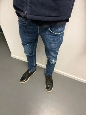 Dondup George  - Säljer ett par blå dondup med slitningar och färgstänk för en trendig, avslappnad look. Jeansen har klassisk femficksdesign och smal passform som sitter snyggt längs benen. Perfekta för dig som gillar streetstyle och vill sticka ut lite extra. Passar större storlekar också