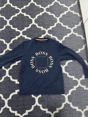 Mörkblå sweatshirt från Hugo Boss - Mörkblå sweatshirt från Hugo Boss med rund halsringning och stort tryck med texten 'BOSS' i vitt på bröstet. Tröjan har långa ärmar och en avslappnad passform, perfekt för en casual look.