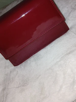 Röd lackad handväska med topphandtag - Snygg röd handväska i glansigt lackmaterial med stilren, fyrkantig form och ett smidigt topphandtag. Perfekt för dig som vill sticka ut med en färgstark accessoar. Väskan har en enkel och modern design utan synliga loggor.