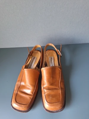 Bruna läderskor med slingback från Marc O'Polo - Snygga bruna skor i äkta läder från Marc O'Polo. Skorna har fyrkantig tå, öppen häl med justerbart spänne och en stilren design. Perfekta för dig som gillar klassiska men unika detaljer. Passar till många olika stilar och är riktigt bekväma.