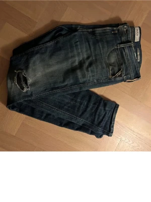Mörkblå slitna jeans från replay - Säljer ett par mörkblå jeans från replay med slitningar och hål vid knät. Klassisk femficksmodell med dragkedja och knapp. Jeansen har en tvättad look och är tillverkade i denim. Perfekta för en avslappnad och trendig stil.