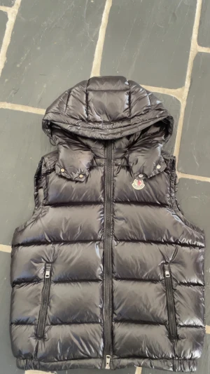 Svart dunväst från Moncler med huva - Snygg svart dunväst från Moncler med glansig finish och avtagbar huva. Västen har två dragkedjefickor fram, quiltad design och Moncler-logga på bröstet. Perfekt för lager-på-lager och streetstyle.