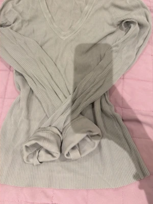 Ljusgrå ribbad långärmad tröja - Säljer en ljusgrå ribbad långärmad tröja med v-ringning. Tröjan har en slim fit passform och är tillverkad i ett mjukt, stretchigt material som sitter snyggt på kroppen. Perfekt för lager på lager och enkel att matcha med olika outfits.