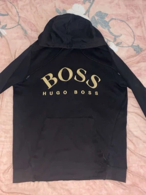 Svart, guld Hugo Boss ( Elimited Edition ) - Säljer en svart hoodie från Hugo Boss med stor guldfärgad BOSS-logga och texten HUGO BOSS på ryggen. Hoodien har huva och en stor magficka framtill. Tillverkad i mjukt material som passar perfekt för en chill och stilren look. Tröjan är elimited och säljs inte mer.