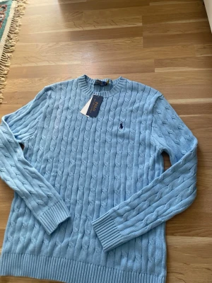 Polo Ralph Lauren Cableknit - Snygg ljusblå kabelstickad tröja från Polo Ralph Lauren med rund halsringning och klassisk broderad logga på bröstet. Tröjan har långa ärmar och ribbade muddar vid ärmslut och nederkant. Perfekt för dig som gillar stilrena och tidlösa plagg.
