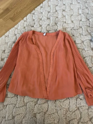Plisserad korallfärgad blus - Säljer en snygg korallfärgad blus med plisserade långärmar och knappar framtill. Blusen har en lös passform och är tillverkad i ett lätt, flowy material som ger en skön känsla. Perfekt att styla med jeans eller kjol för en trendig look.