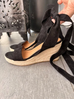 Svarta kilklackade espadrilles med knytband - Snygga svarta espadrilles med kilklack i flätad jute och breda knytband runt ankeln. Ovandelen är i mockaimitation och sulan har en naturlig look. Perfekta för en trendig och avslappnad stil.