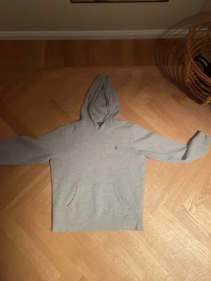 Grå hoodie från Polo Ralph Lauren - Säljer en stilren grå hoodie från Polo Ralph Lauren med klassisk känguruficka och liten broderad logga på bröstet. Hoodien har en mjuk bomullskänsla och en skön huva. Perfekt för chill dagar eller när du vill ha en clean look. Inga defekter. Storlek 164cm 14 år
