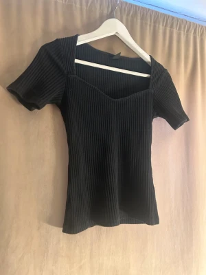 Svart ribbad topp från Gina Tricot - Snygg svart ribbad topp från Gina Tricot med korta ärmar och v-ringad hals. Toppen har en figurnära passform och är tillverkad i ett stretchigt material som sitter bekvämt. Perfekt att styla med jeans eller kjol.