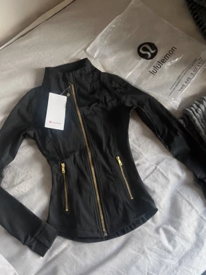 Svart jacka från Lululemon med guldzip - Snygg svart jacka från Lululemon med figurnära passform och guldfärgade dragkedjor framtill och på fickorna. Tillverkad i stretchigt syntetmaterial och har hög krage samt långa ärmar. Perfekt för en sportig och stilren look.