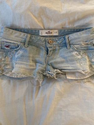 Ljusblå  jeansshorts från Hollister - Säljer ett par ljusblå jeansshorts från Hollister med slitna detaljer och rå kant. Klassisk femficksmodell med knapp och dragkedja framtill. Perfekta för varma dagar och ger en avslappnad vibe💕💕