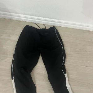 Svarta mjukisbyxor från BOSS - Säljer ett par svarta mjukisbyxor från BOSS med vita detaljer längs sidorna och vita dragkedjor vid fickorna. Byxorna har dragsko i midjan och är tillverkade i mjukt material för maximal komfort. Perfekta för en avslappnad stil.