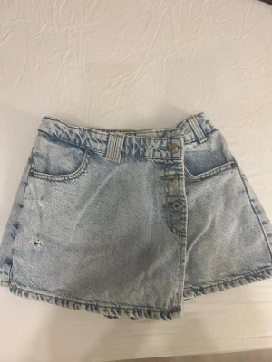 Ljusblå denimskort från Zara - Säljer ett par ljusblå denimskorts från Zara med cool wrap-front och slitna detaljer. Klassisk femficksmodell, bälteshällor och rå kant nedtill. Perfekt för dig som gillar en avslappnad och trendig stil. Har små tecken på användning på knapparna på bild fem och en väldigt ljus gul fläck på baksidan vilket inte syns så bra på bild fyra.