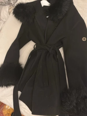 Svart kappa med fuskpälsdetaljer - Stilren svart kappa med stora fuskpälsdetaljer vid krage och ärmslut. Knytskärp i midjan och diskret logga på ärmen. Perfekt för dig som vill ha en trendig och elegant look under kalla dagar.
