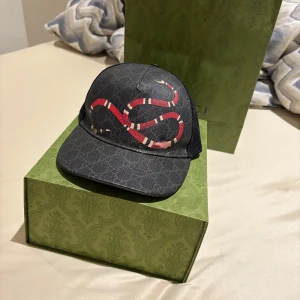 Gucci keps med orm och GG-mönster - Äkta…..Snygg svart keps från Gucci med klassiskt GG-mönster och en röd orm på framsidan. Baksidan har mesh och justerbart band med röd och marinblå rand. Perfekt statement-piece för dig som vill sticka ut. Materialmix med bomull och polyester.