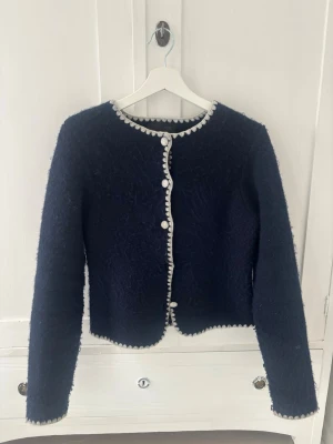 Mörkblå kofta från Zara med vita detaljer - Säljer en mörkblå kofta från Zara med rund halsringning och dekorativa vita kanter samt vita klädda knappar framtill. Koftan är stickad i ett fluffigt material och har långa ärmar.använd fatals gånger🩷