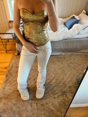 Guldig axelbandslös korsett - Säljer en snygg axelbandslös korsett i metallic guld med strukturmönster. Korsetten har en hjärtformad urringning och markerad midja. Perfekt för att lyfta vilken outfit som helst med sin glansiga finish.