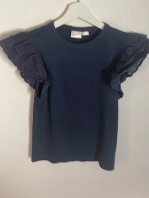 Mörkblå topp med volangärmar - Supersnygg mörkblå topp med volangdetaljer på ärmarna. Rund halsringning och korta, vågiga ärmar som ger en unik look. Perfekt att styla med jeans eller kjol för en trendig outfit.