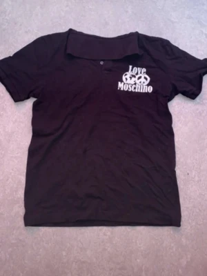 Svart Love Moschino skjorta - Säljer en svart skjorta från Love Moschino med vit logga och tryck på bröstet. Skjortan har klassisk krage, kort ärm och knappar framtill. Perfekt för dig som gillar streetwear och vill ha något från ett känt märke.