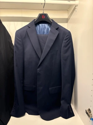 Mörkblå kavaj från Dressmann - Snygg mörkblå kavaj från Dressmann i slim fit-modell. Klassisk design med två framfickor med lock, bröstficka och knappar vid ärmslut. Perfekt för dig som vill ha en stilren och modern look. Kavaj i storlek
