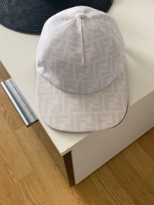 Beige keps från Fendi med FF-mönster - Säljer en beige keps från Fendi med det klassiska FF-logomönstret över hela kepsen. Kepsen har böjd skärm och är tillverkad i ett slitstarkt tyg. Perfekt för dig som vill ha en stilren och exklusiv accessoar till din outfit.