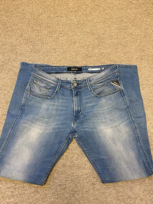 Blå Replay Anbass jeans  - Snygga blå jeans från Replay som har ljusa slitningar framtill och baktill. Jeansen  har normal passform, bälteshällor och knappgylf. Perfekta för sommaren och ger en avslappnad look.