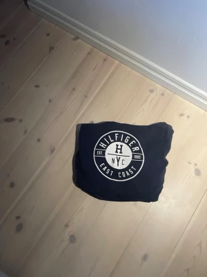 Mörkblå Hilfiger hoodie - Snygg mörkblå hoodie från Hilfiger med stort vitt NYC-tryck på bröstet och röda snören i huvan. Insidan av huvan är grå och tröjan har ribbade muddar vid ärmslut och nederkant. Perfekt för en avslappnad streetstil.