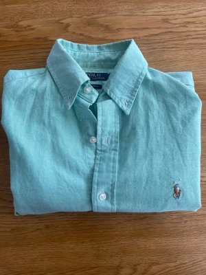 Ljusgrön skjorta från Polo Ralph Lauren - Klassisk ljusgrön skjorta från Polo Ralph Lauren med button-down krage och broderad logga på bröstet. Skjortan har vita knappar och är tillverkad i mjuk bomull. Perfekt för en stilren och avslappnad look.
