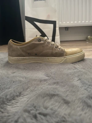 Beige sneakers från Lanvin i mocka - Snygga beige sneakers från Lanvin med ovandel i mocka och tåparti i blankt material. Klassisk låg modell med beiga snören och gummisula. Perfekta för dig som gillar stilrena och exklusiva sneakers. Pris kan diskuteras vid snabb affär,        