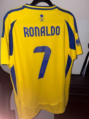 Al Nassr Ronaldo matchtröja gul/blå - Gul och blå Al Nassr matchtröja från Adidas med Ronaldo och nummer 7 på ryggen. Tröjan har klubbmärke, sponsorloggor och blå detaljer längs sidor och axlar. Tillverkad i lätt och ventilerande material, perfekt för fotbollsträning eller match.