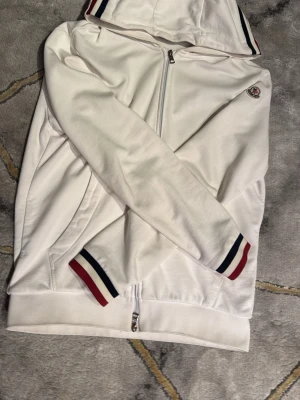 Vit Moncler hoodie med ränder - Vit hoodie från Moncler med dragkedja, huva och ribbade muddar. Detaljer i form av röda och marinblå ränder på huva och ärmslut samt Moncler-logga på bröstet. Tillverkad i mjukt bomullsmaterial för en clean och sportig look.