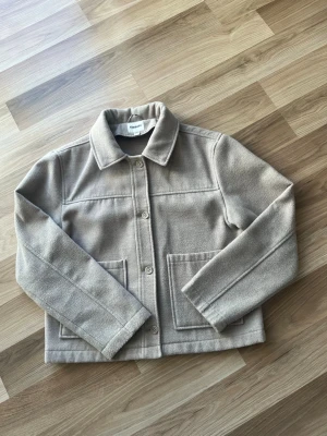 Beige kappa från Kappahl - Kappa i beige filtmaterial från Kappahl med rak passform, klassisk krage och stora fyrkantiga fickor framtill. Stängs med fyra knappar och har markerade sömmar för en clean look. Perfekt för lager-på-lager och enkel att styla.