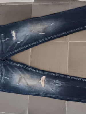 Mörkblå slitna jeans med hål - Säljer ett par mörkblå jeans med slitningar och hål på benen. Jeansen har klassisk femficksdesign, raka ben och snygga slitdetaljer både fram och bak. Perfekta för en avslappnad och trendig look.