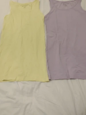Pastellgula och lila linnetoppar - Två stilrena linnetoppar i pastellgul och ljuslila. Båda är ärmlösa med klassisk rund halsringning och raka linjer, perfekta att matcha med jeans eller kjol. Materialet känns mjukt och lätt, vilket gör dem sköna att bära under varma dagar. Storlek 146/152