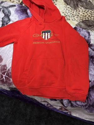 Röd hoodie från GANT med tryck - Snygg röd hoodie från GANT med broderad logga och texten 'American Sportswear' på bröstet. Klassisk känguruficka framtill och ribbade muddar. Perfekt för en sportig och avslappnad stil. Materialet är mjukt och bekvämt.