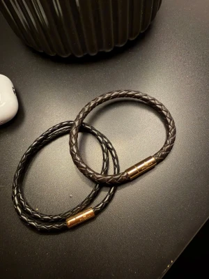 Svarta och bruna flätade armband - Två stilrena armband från Skultuna i svart och mörkbrunt flätat läder med guldfärgade magnetlås. Perfekta accessoarer för dig som gillar en clean och modern look. Passar både till klocka och ensamt för en snygg detalj. Utan box.